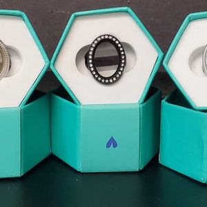 May include: Five teal hexagonal boxes with white lids, each containing a silver locket with a different design. The lockets have a round, oval-shaped opening with a mirror, black, or silver background.