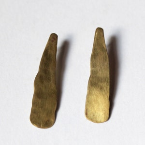 Puede incluir: Un par de pendientes de color dorado con una forma irregular texturizada. Los pendientes están hechos de metal y tienen un acabado ligeramente martillado.