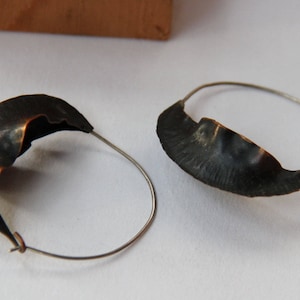 May include: A pair of hoop earrings with a unique, asymmetrical design. The hoops are made of silver and feature a textured, copper-coloured metal piece that is shaped like a crescent moon.