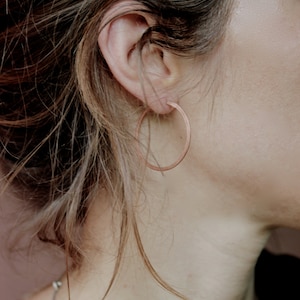 Könnte beinhalten: Nahaufnahme eines Ohrs mit einem schlichten, dünnen, kupfernen Creolenohrring. Der Ohrring ist im klassischen, minimalistischen Stil gehalten.