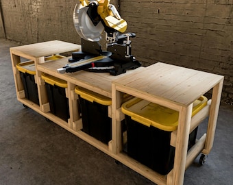Può includere: Un banco da lavoro in legno con una sega a smusso e quattro contenitori di stoccaggio. Il banco da lavoro ha ruote ed è realizzato in legno grezzo. I contenitori di stoccaggio sono neri con coperchi gialli.