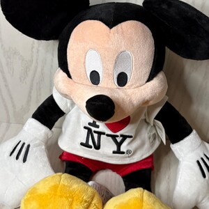 May include: A plush Mickey Mouse doll with black ears, black arms, and white gloves. The doll wears a white shirt with "I &hearts; NY" printed on it, red shorts, and yellow shoes. The doll is a popular character.