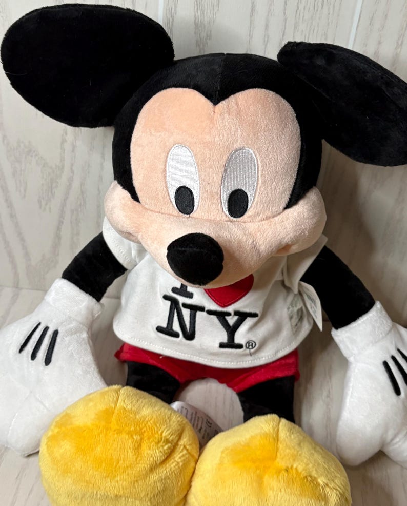 May include: A plush Mickey Mouse doll with black ears, black arms, and white gloves. The doll wears a white shirt with "I &hearts; NY" printed on it, red shorts, and yellow shoes. The doll is a popular character.