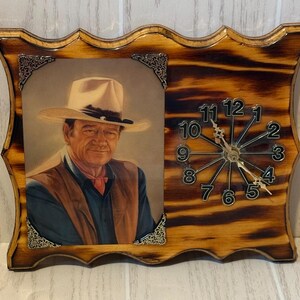 May include: A rectangular wall clock with a wavy edge, featuring a portrait of a man in a cowboy hat on the left and a clock face with black numbers and silver hands on the right. The frame is made of wood with a dark, scorched finish.