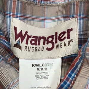 May include: A close-up of a Wrangler Rugged Wear shirt tag. The tag is white with brown text and a brown logo. The tag says "Wrangler Rugged Wear" and "RWL68T M/M/M 100% COTTON 100% ALGODON 100% COTON MADE IN BANGLADESH".