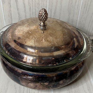 May include: A vintage silver-plated serving dish with a glass insert. The dish has a domed lid with a decorative finial and ornate handles. The silver finish shows signs of age, adding to its antique charm. The dish is likely used for serving food.