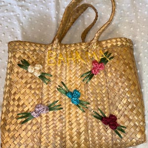 May include: Woven straw tote bag with the word "BAHAMA" embroidered in yellow. The bag is decorated with colorful floral accents in white, pink, blue, purple, and red. The bag has woven handles.