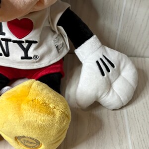 May include: A plush Mickey Mouse doll wearing a white t-shirt with a red heart and the text "I NY". The doll has black arms, red shorts, and yellow shoes. The white gloves have three black lines.