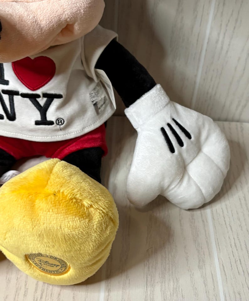 May include: A plush Mickey Mouse doll wearing a white t-shirt with a red heart and the text "I NY". The doll has black arms, red shorts, and yellow shoes. The white gloves have three black lines.