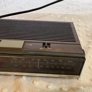 Puede incluir: Un radio reloj marr&oacute;n vintage con un cable de alimentaci&oacute;n negro. La radio tiene un sintonizador FM/AM, una funci&oacute;n de repetici&oacute;n y una bater&iacute;a de respaldo. La cara muestra las marcas de frecuencia en MHz y kHz.