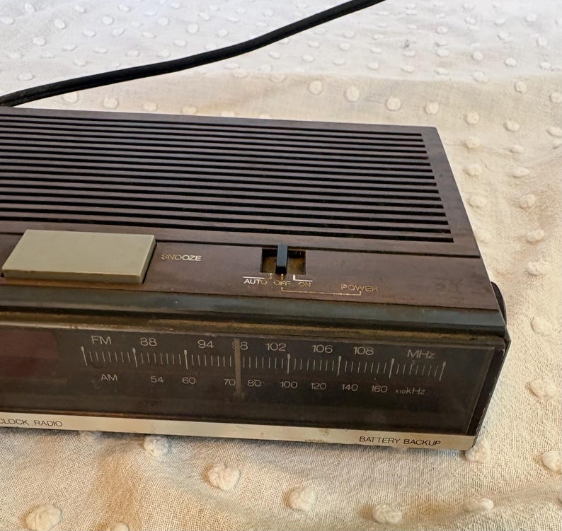 Puede incluir: Un radio reloj marr&oacute;n vintage con un cable de alimentaci&oacute;n negro. La radio tiene un sintonizador FM/AM, una funci&oacute;n de repetici&oacute;n y una bater&iacute;a de respaldo. La cara muestra las marcas de frecuencia en MHz y kHz.
