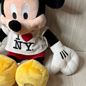 May include: A plush Mickey Mouse doll wearing a white t-shirt with "I ❤️ NY" printed on it, red shorts, and yellow shoes. The doll has black ears, a black nose, and white gloves with three black lines.