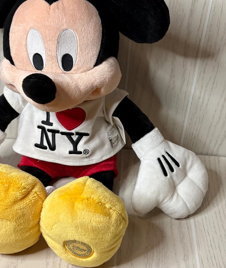 May include: A plush Mickey Mouse doll wearing a white t-shirt with "I ❤️ NY" printed on it, red shorts, and yellow shoes. The doll has black ears, a black nose, and white gloves with three black lines.