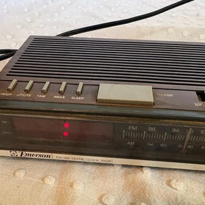 Puede incluir: Una radio reloj digital FM/AM Emerson vintage. La radio tiene una parte superior marr&oacute;n oscuro con l&iacute;neas horizontales y una pantalla digital. Los botones incluyen Hora, Minuto, Tiempo, Despertar, Dormir y Repetici&oacute;n. La radio tiene un dial AM/FM.