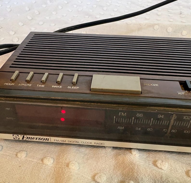 Puede incluir: Una radio reloj digital FM/AM Emerson vintage. La radio tiene una parte superior marr&oacute;n oscuro con l&iacute;neas horizontales y una pantalla digital. Los botones incluyen Hora, Minuto, Tiempo, Despertar, Dormir y Repetici&oacute;n. La radio tiene un dial AM/FM.