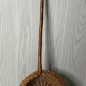 May include: A long-handled, round, woven wicker basket with a deep, circular bowl and a sturdy, slender handle extending from the top rim. The basket features a natural light brown finish with a braided edge detail around the circular opening.