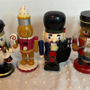 May include: Four decorative wooden nutcrackers in various designs. One is a gingerbread man holding a lollipop, and others are dressed in military-style uniforms. Each has a unique hat and facial expression, with detailed paintwork and accessories.