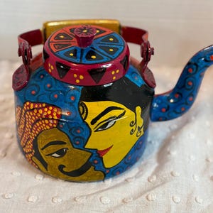 May include: A hand-painted, decorative teapot with a vibrant design. The teapot features a blue body with painted faces, a red lid, and a blue spout. The handle and lid handle are red. The design includes yellow, red, and black accents.