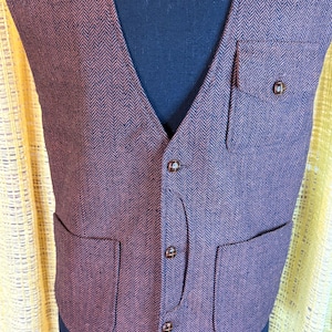 May include: A brown and burgundy herringbone vest with a V-neck, two front pockets, and a chest pocket. The vest has brown buttons and is displayed on a black mannequin.
