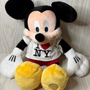 May include: A plush Mickey Mouse doll wearing a white t-shirt with "I ❤️ NY" printed on it. The doll has black ears, a pink face, white gloves, red shorts, and two yellow plush circles. A Disney logo is on one of the yellow circles.