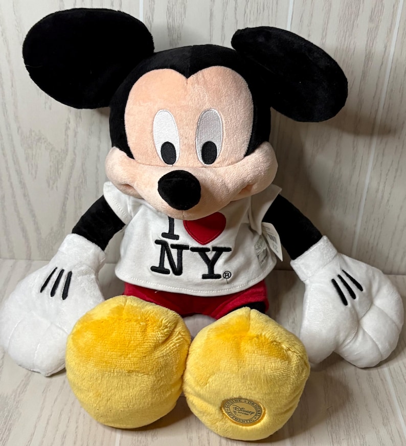 May include: A plush Mickey Mouse doll wearing a white t-shirt with "I ❤️ NY" printed on it. The doll has black ears, a pink face, white gloves, red shorts, and two yellow plush circles. A Disney logo is on one of the yellow circles.
