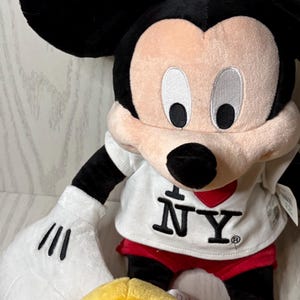 May include: A plush Mickey Mouse doll with black ears, arms, and legs. The doll has a white shirt with "I ❤️ NY" printed on it, red shorts, and yellow shoes. The face is pink with black eyes and a black nose.