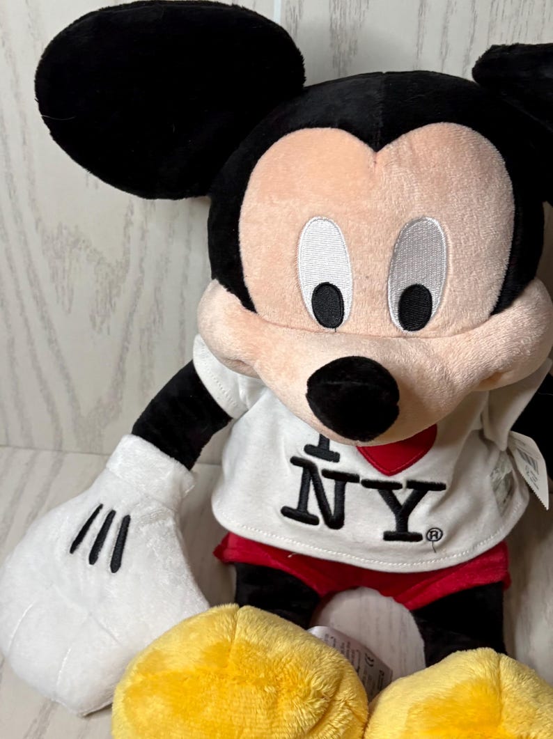 May include: A plush Mickey Mouse doll with black ears, arms, and legs. The doll has a white shirt with "I ❤️ NY" printed on it, red shorts, and yellow shoes. The face is pink with black eyes and a black nose.