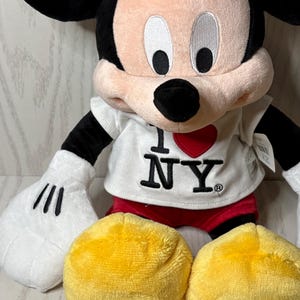 May include: A plush Mickey Mouse doll wearing a white t-shirt with "I ❤️ NY" printed on it. The doll has black ears, arms, and a tail, with white gloves and red shorts. Two yellow plush circles are in front of the doll.