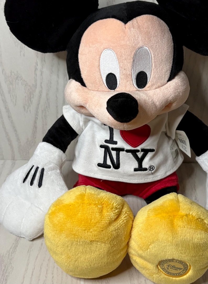 May include: A plush Mickey Mouse doll wearing a white t-shirt with "I ❤️ NY" printed on it. The doll has black ears, arms, and a tail, with white gloves and red shorts. Two yellow plush circles are in front of the doll.