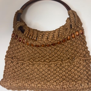 May include: A brown crocheted handbag with a dark brown wooden handle. The bag is adorned with small wooden beads along the top edge. The bag's texture is visible, showcasing the intricate crochet work. The bag is designed for carrying items.