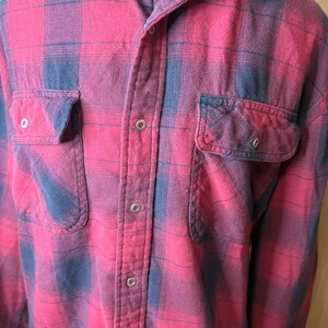 May include: A red and navy blue plaid flannel shirt. The shirt has two buttoned chest pockets and a button-down front. The collar is buttoned, and the shirt appears to be made of a soft, textured fabric.