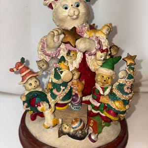 May include: A whimsical Christmas figurine featuring a white cat dressed as Mrs. Santa Claus, surrounded by several cat figurines. The figurine is adorned with festive colors, including red, green, and gold, and includes a Christmas tree and star. The base reads "Mrs. Santa Claws."