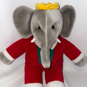 May include: A gray plush elephant wearing a red suit with a green shirt and a yellow crown. The elephant has large ears and a long trunk.