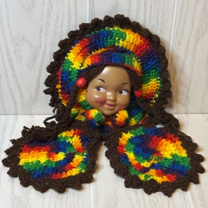 May include: A doll head with brown hair and red beads is framed by a crocheted rainbow-colored headdress. The headdress features a dark brown scalloped border and two long, matching crocheted rainbow-colored extensions with dark brown edges.