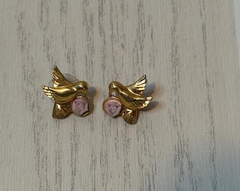 May include: A pair of gold-toned stud earrings shaped like hummingbirds, each featuring a small, light pink sculpted rose accent positioned near the bird's chest. The earrings have a metallic finish and detailed wing and tail feather textures.