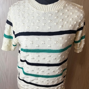 May include: Cream-colored short-sleeve sweater with textured bobble stitch detailing. The sweater features horizontal stripes in navy and teal. The neckline and sleeve cuffs are ribbed.