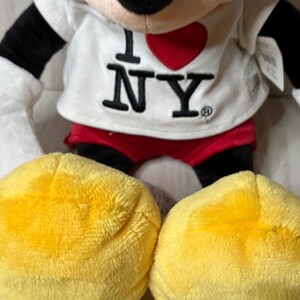 May include: A plush Mickey Mouse doll wearing a white shirt with "I ❤️ NY" printed on it. The doll has black ears, arms, and a red shorts. The large yellow feet are visible in the foreground.