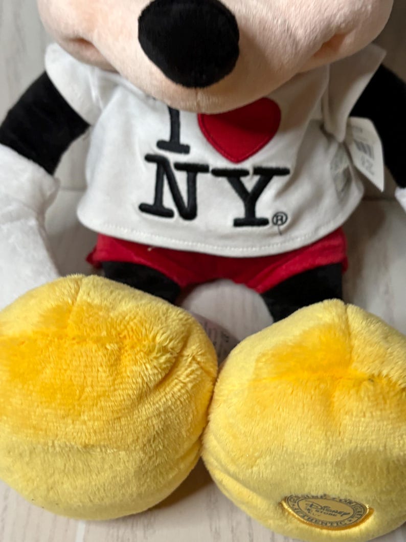 May include: A plush Mickey Mouse doll wearing a white shirt with "I ❤️ NY" printed on it. The doll has black ears, arms, and a red shorts. The large yellow feet are visible in the foreground.