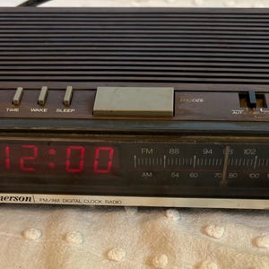 Puede incluir: Un radio reloj digital FM/AM Emerson vintage con acabado de veta de madera marr&oacute;n oscuro. La pantalla digital roja muestra la hora como 12:00. Incluye botones de repetici&oacute;n, hora, despertador y dormir. La radio tiene un sintonizador AM/FM.