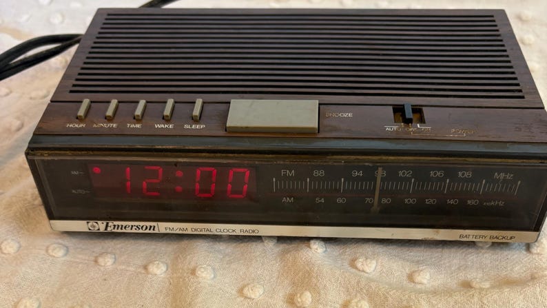 Puede incluir: Un radio reloj digital FM/AM Emerson vintage con acabado de veta de madera marr&oacute;n oscuro. La pantalla digital roja muestra la hora como 12:00. Incluye botones de repetici&oacute;n, hora, despertador y dormir. La radio tiene un sintonizador AM/FM.