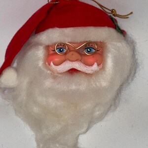 May include: A vintage Santa Claus head ornament. The ornament features a red hat, a white beard, and gold wire-rimmed glasses. The face is painted with rosy cheeks and blue eyes. A gold string is attached for hanging.