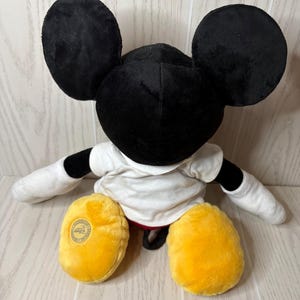 May include: A plush Mickey Mouse doll, featuring large black ears, a black head, and a white shirt. The doll has yellow feet and white gloves. The doll is sitting on a light-colored wooden surface.