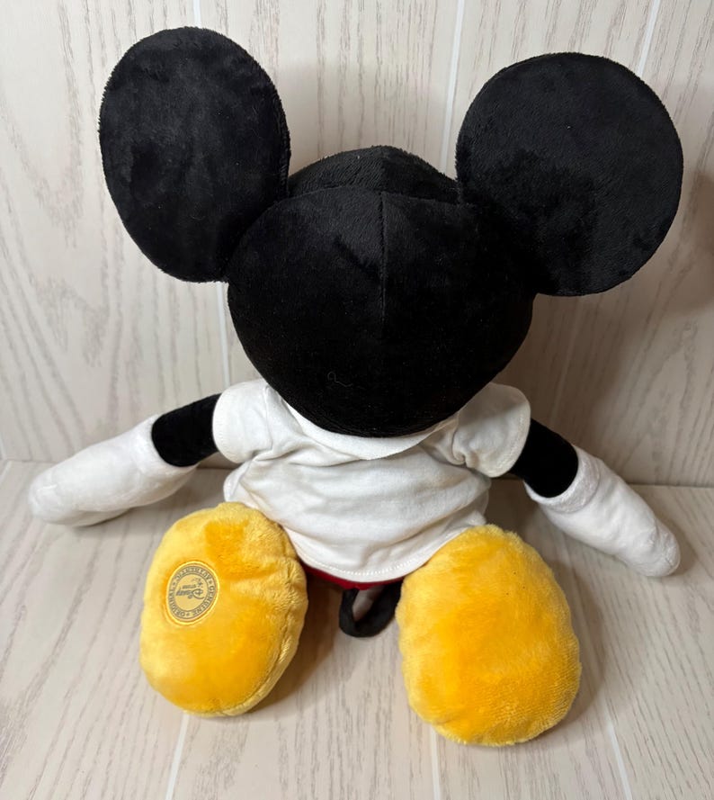 May include: A plush Mickey Mouse doll, featuring large black ears, a black head, and a white shirt. The doll has yellow feet and white gloves. The doll is sitting on a light-colored wooden surface.