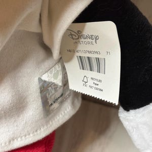 May include: A close-up of a Disney Store tag attached to a plush toy. The tag is white with the Disney Store logo and product information, including a barcode and the text "RECYCLED Paper". The plush toy has red, black, and white elements.