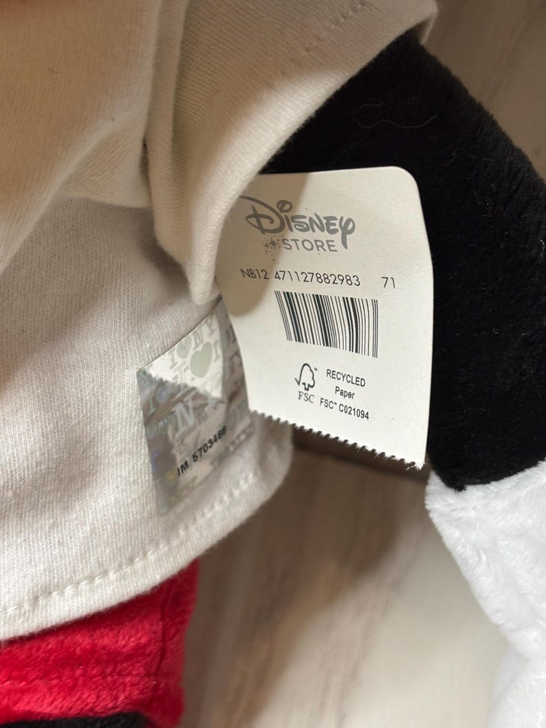 May include: A close-up of a Disney Store tag attached to a plush toy. The tag is white with the Disney Store logo and product information, including a barcode and the text "RECYCLED Paper". The plush toy has red, black, and white elements.