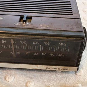 Puede incluir: Primer plano de un reloj despertador digital marr&oacute;n vintage. La esfera del reloj muestra n&uacute;meros de frecuencia de radio y el texto "BATTERY BACKUP". La parte superior tiene un bot&oacute;n "SNOOZE" y un interruptor "AUTO OFF ON".