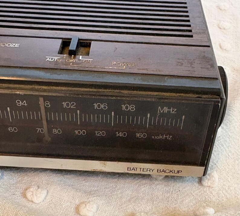 Puede incluir: Primer plano de un reloj despertador digital marr&oacute;n vintage. La esfera del reloj muestra n&uacute;meros de frecuencia de radio y el texto "BATTERY BACKUP". La parte superior tiene un bot&oacute;n "SNOOZE" y un interruptor "AUTO OFF ON".