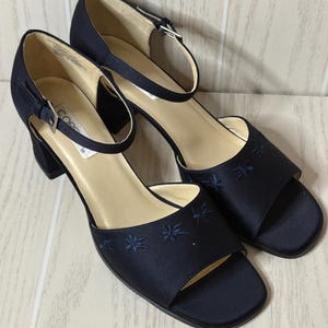 May include: A pair of navy blue satin open-toe sandals with a block heel and an adjustable ankle strap featuring a silver-tone buckle. The front of each shoe is decorated with small, tonal embroidered floral motifs and a light beige inner lining.