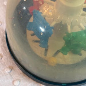 May include: A vintage toy with a clear plastic dome, featuring colorful plastic figures and the word "Go". The base is dark blue, and the figures include blue, green, and red elements. The toy appears to be a spinning or rolling type.