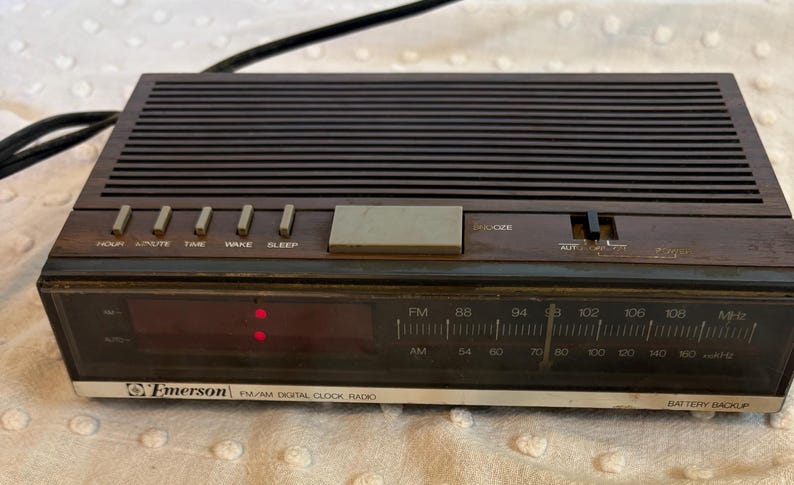 Puede incluir: Un radio reloj digital PM/AM Emerson vintage con un acabado de madera marr&oacute;n oscuro. Incluye una pantalla digital, un dial de frecuencia de radio y botones para hora, minuto, despertar y dormir. El reloj tiene una bater&iacute;a de respaldo.
